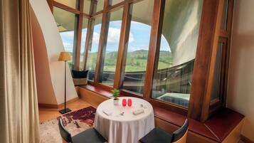 A room with a round table, two chairs, a potted plant, and a view of green hills through large windows.