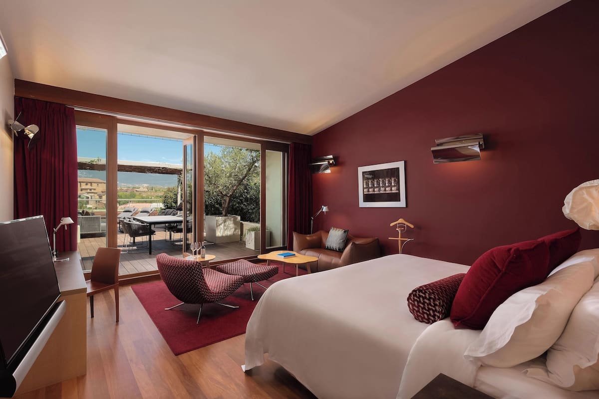 A modern hotel room with a large bed, a TV, a desk, and a balcony with outdoor furniture.