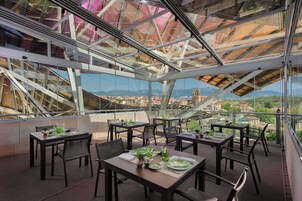 Uma área de refeições no terraço, com mesas, cadeiras, grandes janelas de vidro e vista para a cidade.