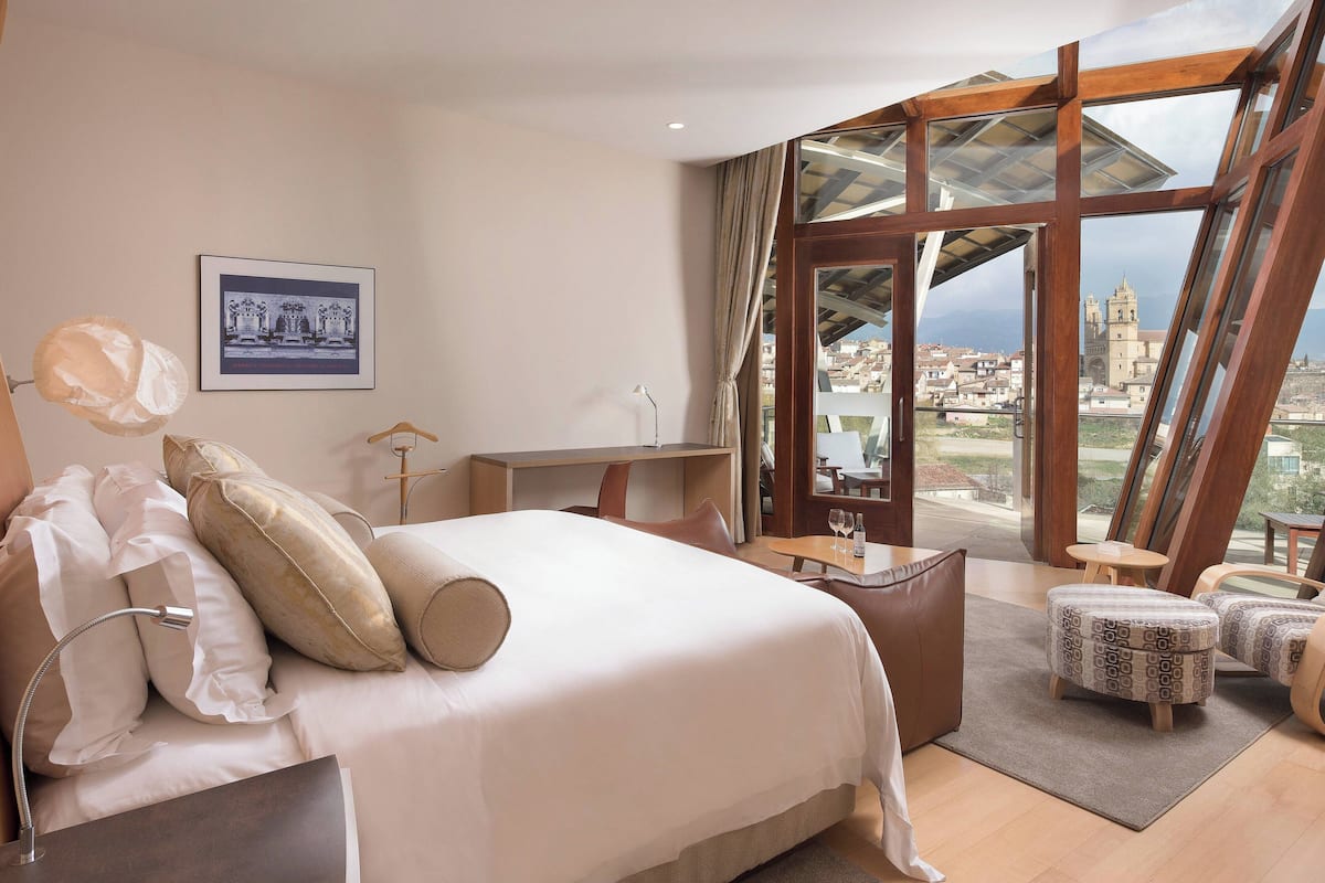 A hotel room with a large bed, a desk, a chair, and a view of a cityscape through large windows.