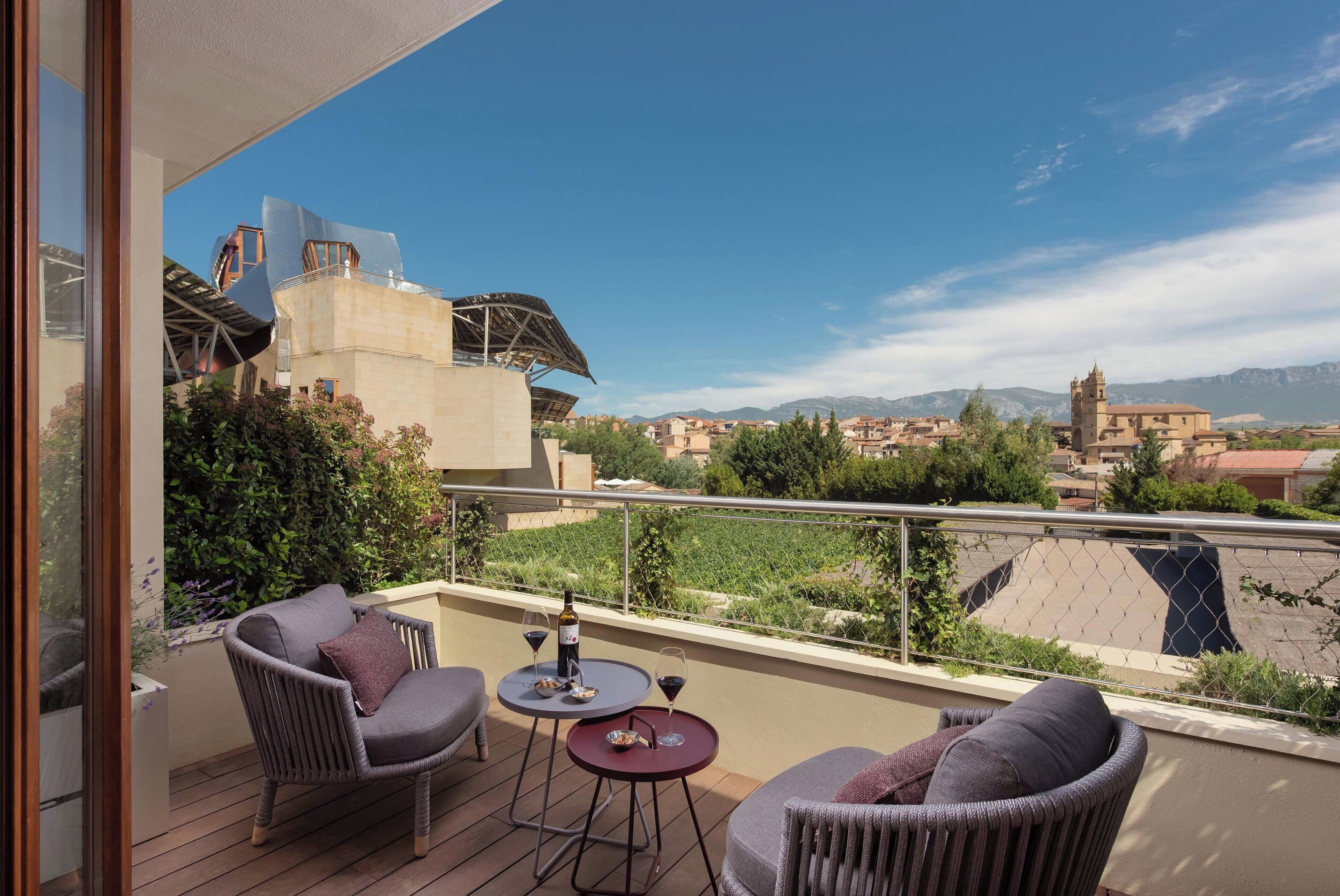 Uma varanda com móveis de vime, uma mesa com copos de vinho e uma garrafa, e vista para uma paisagem urbana com montanhas ao fundo.
