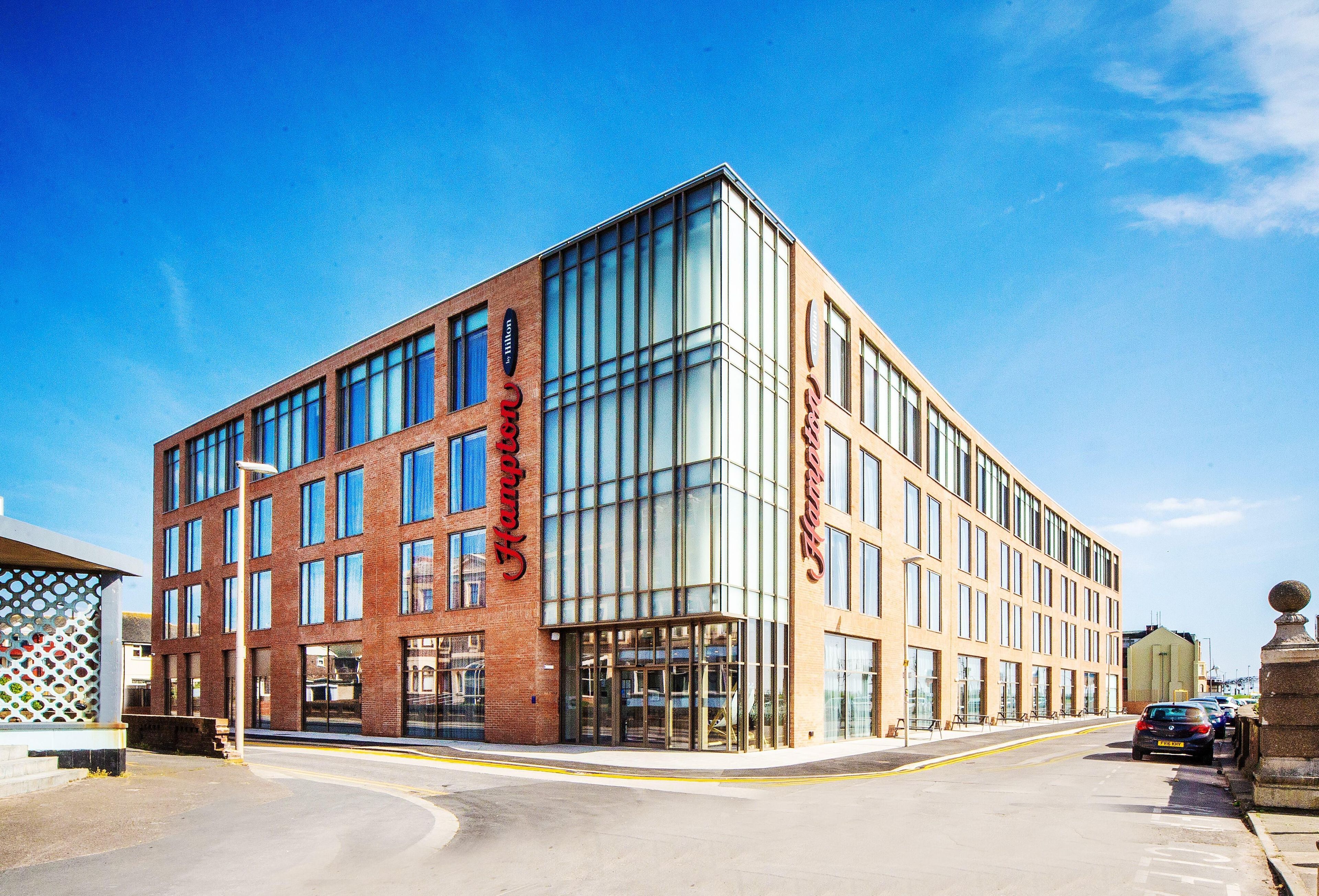 A modern hotel building with a corner entrance, large windows, and a prominent brand name displayed on the facade.