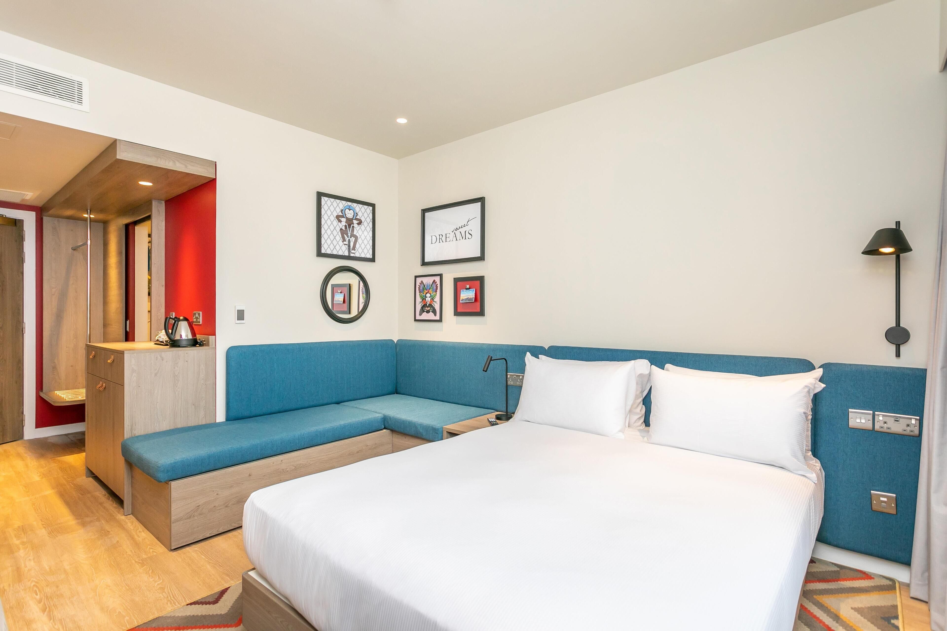 A modern hotel room with a large bed, a blue sofa, a small desk, and framed pictures on the wall.