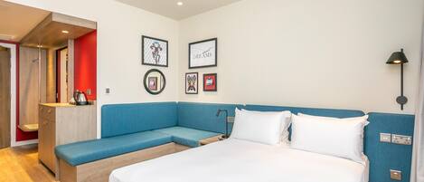 A modern hotel room with a large bed, a blue sofa, a small desk, and framed pictures on the wall.