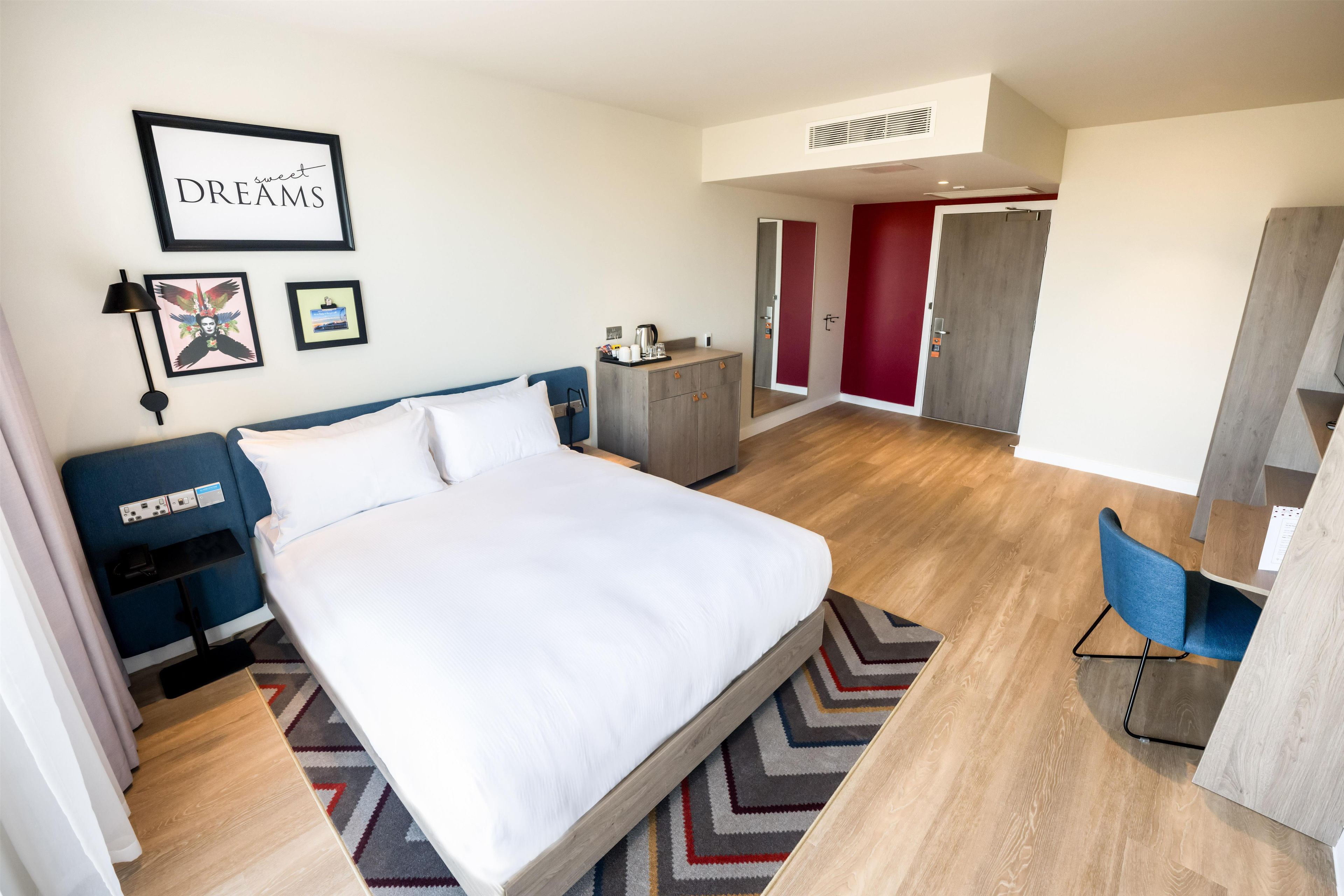 A modern hotel room with a large bed, a desk, a wardrobe, and a wall with framed pictures.