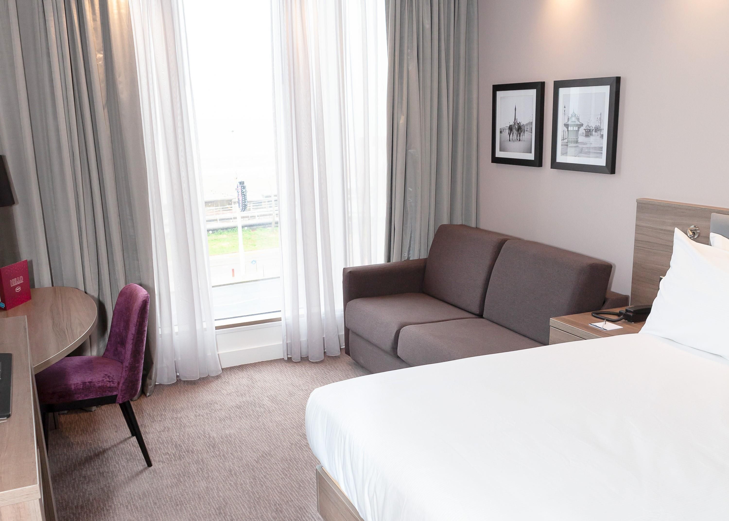 Habitación de hotel con cama, sofá, silla, escritorio y ventana con cortinas. Hay dos cuadros en la pared.