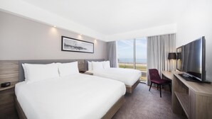 A hotel room with two beds, a desk with a TV, a chair, and a large window with a view of the beach. - Hampton by Hilton Blackpool (Blackpool)