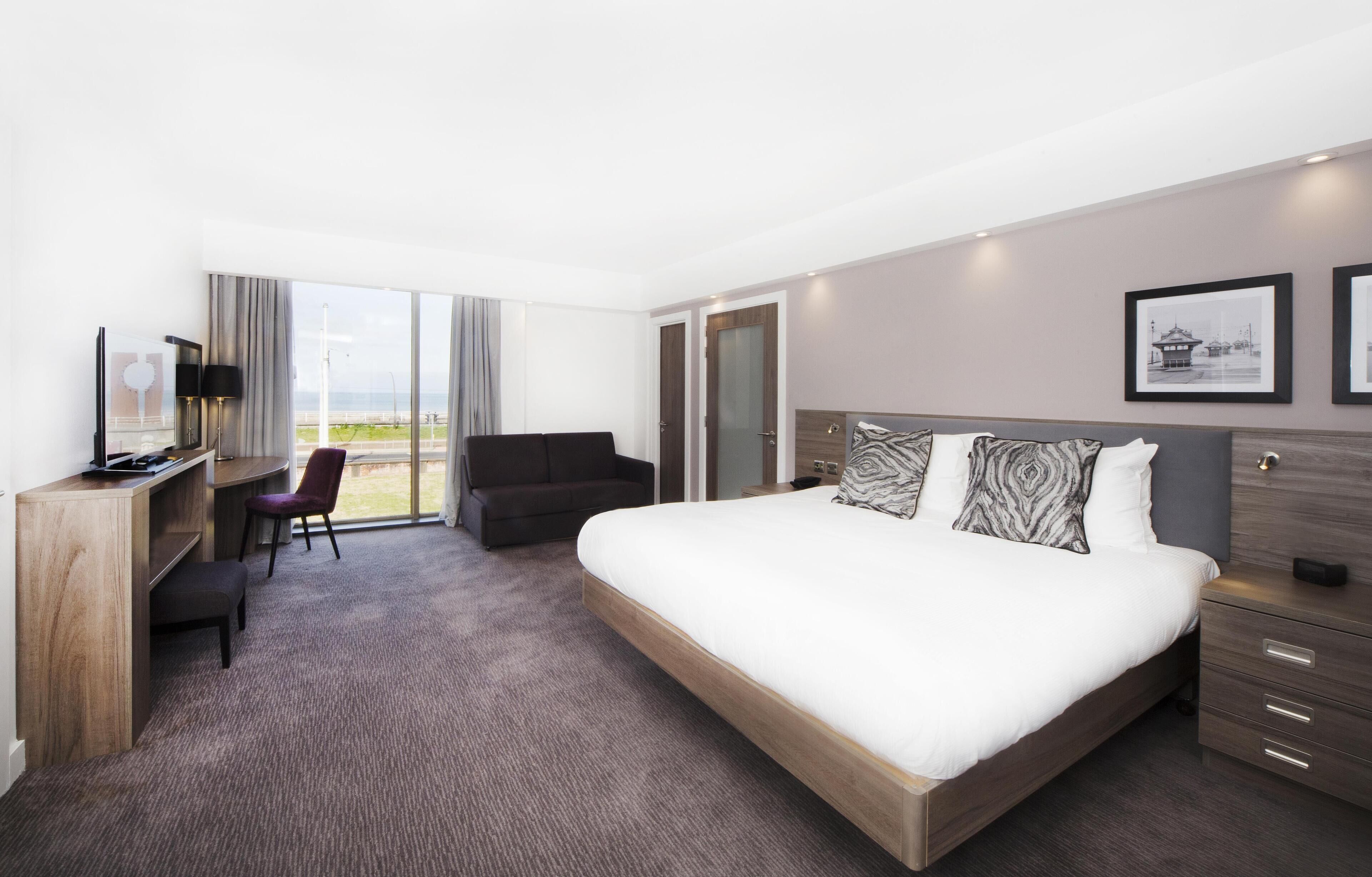 A modern hotel room with a large bed, a desk, a chair, and a view of the outdoors.