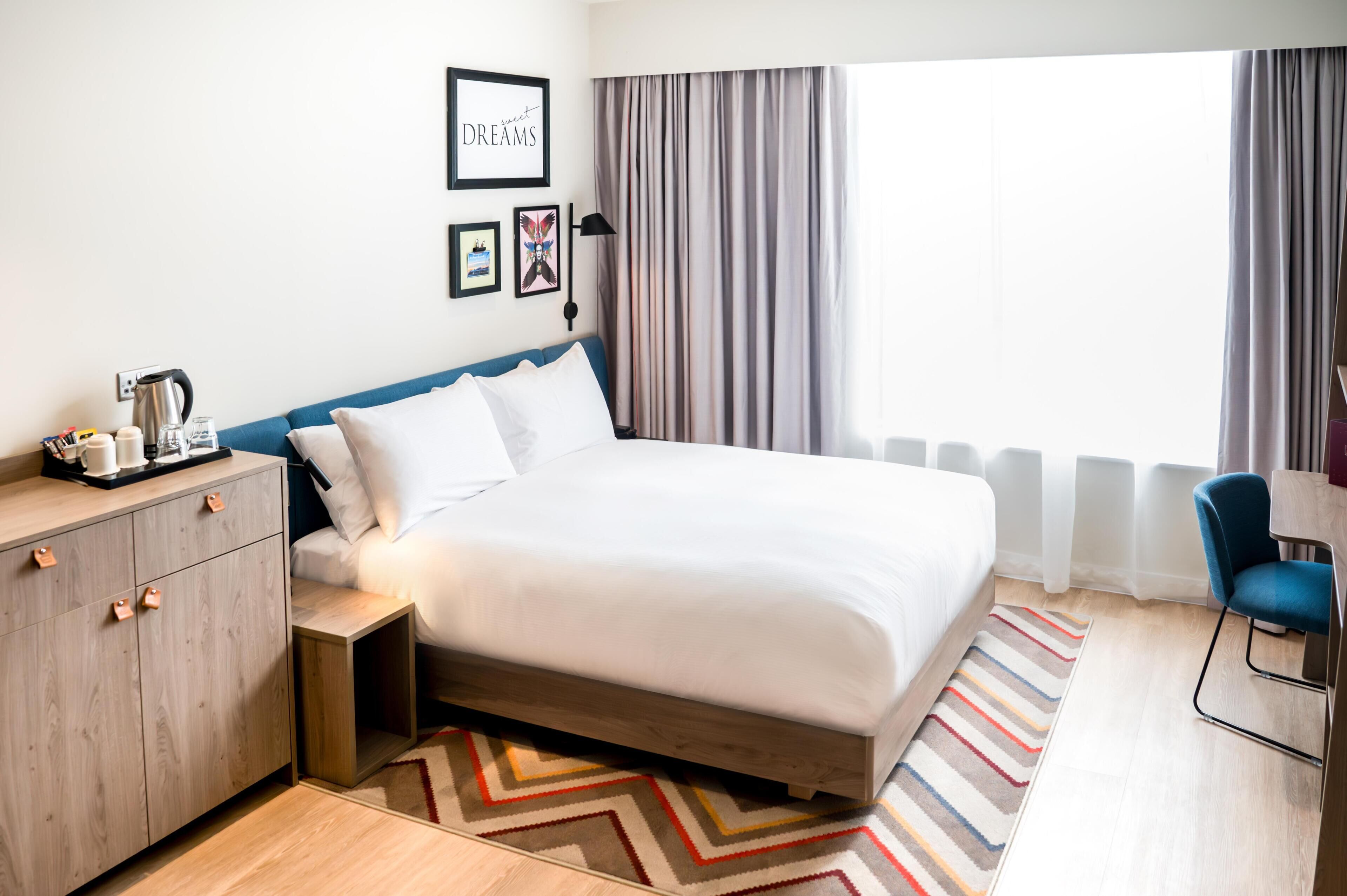 A neatly arranged hotel room with a large bed, a bedside table with a kettle, a small cabinet, a chair, and a window with curtains.