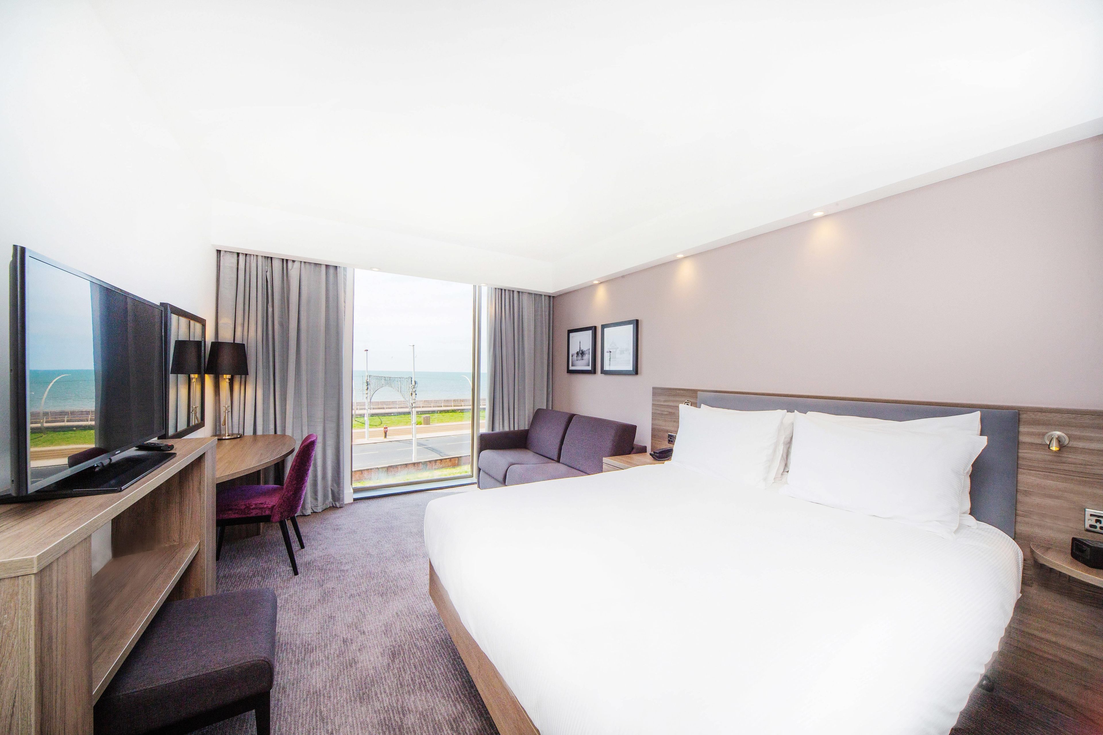 Ein Hotelzimmer mit einem großen Bett, einem Schreibtisch mit Fernseher, einem Sessel und Blick ins Freie.