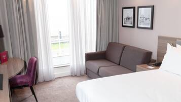 A hotel room with a bed, a sofa, a chair, a desk, and a window with curtains. There are two framed pictures on the wall.