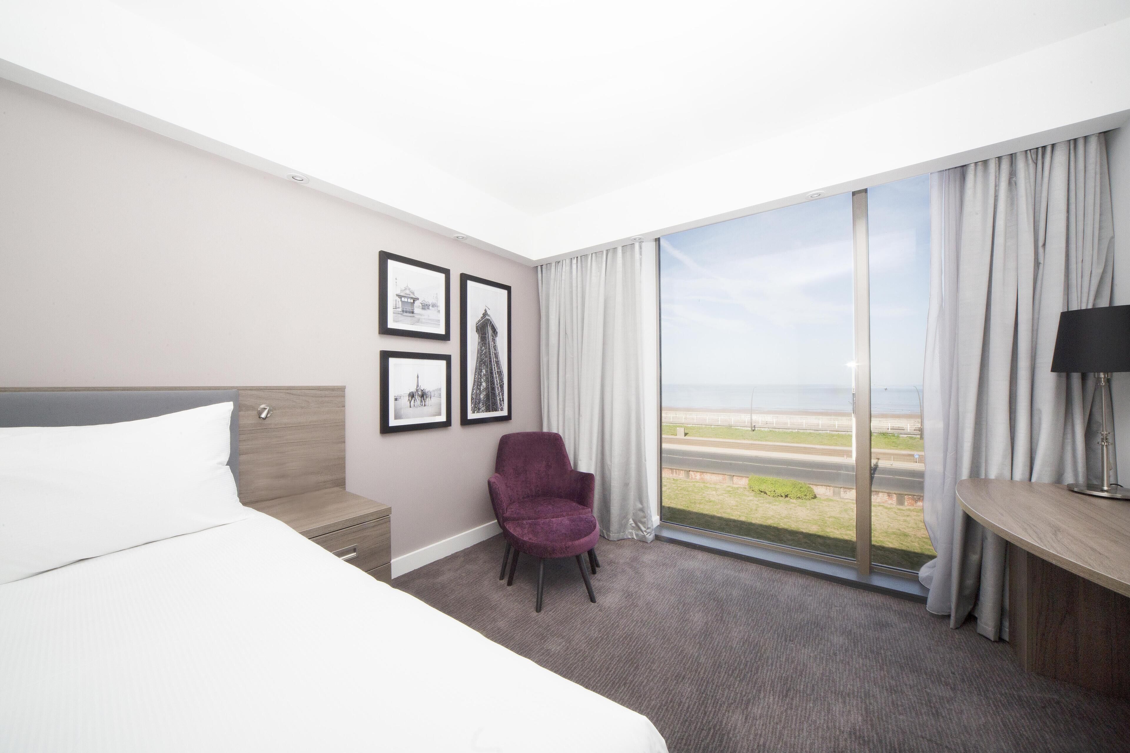 A hotel room with a bed, a chair, a desk, a lamp, and a large window with a view of the beach.
