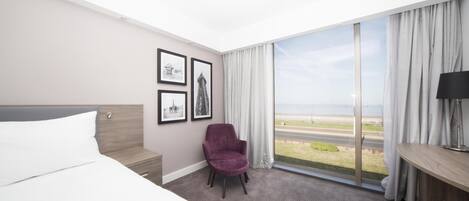 Habitación de hotel con cama, una silla, un escritorio, una lámpara y un ventanal con vista a la playa.