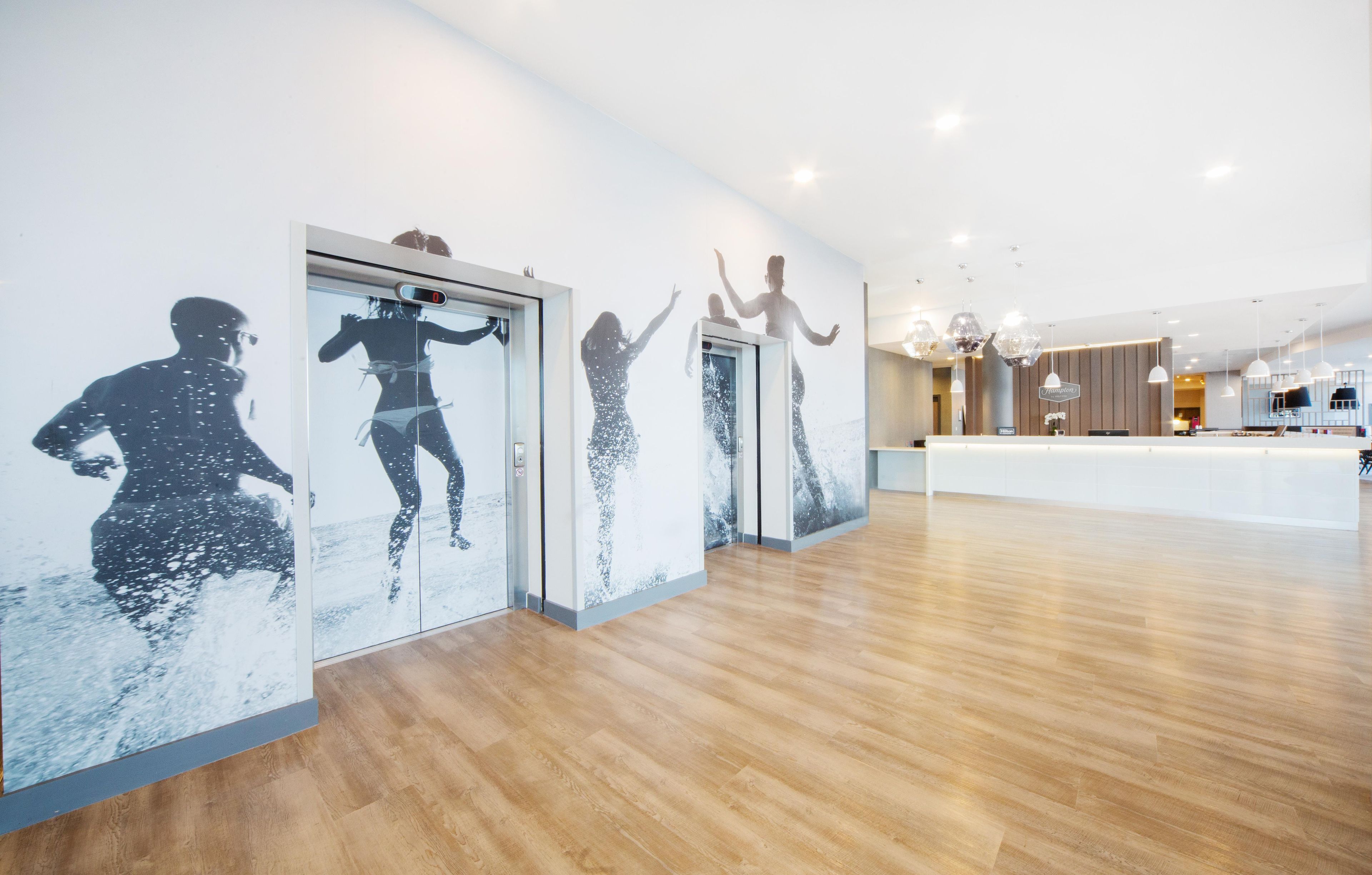 A modern hotel lobby with wooden flooring, a reception desk, and wall art depicting people in motion.