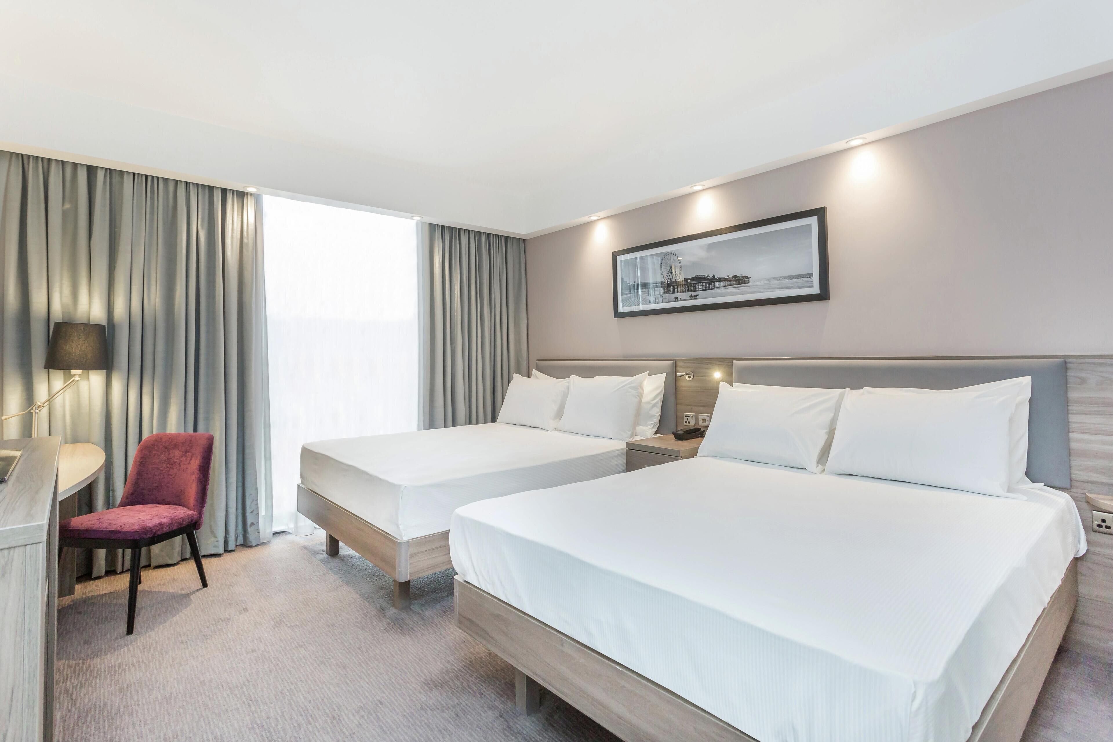 A hotel room with two beds, a desk, a chair, and a large window with curtains.