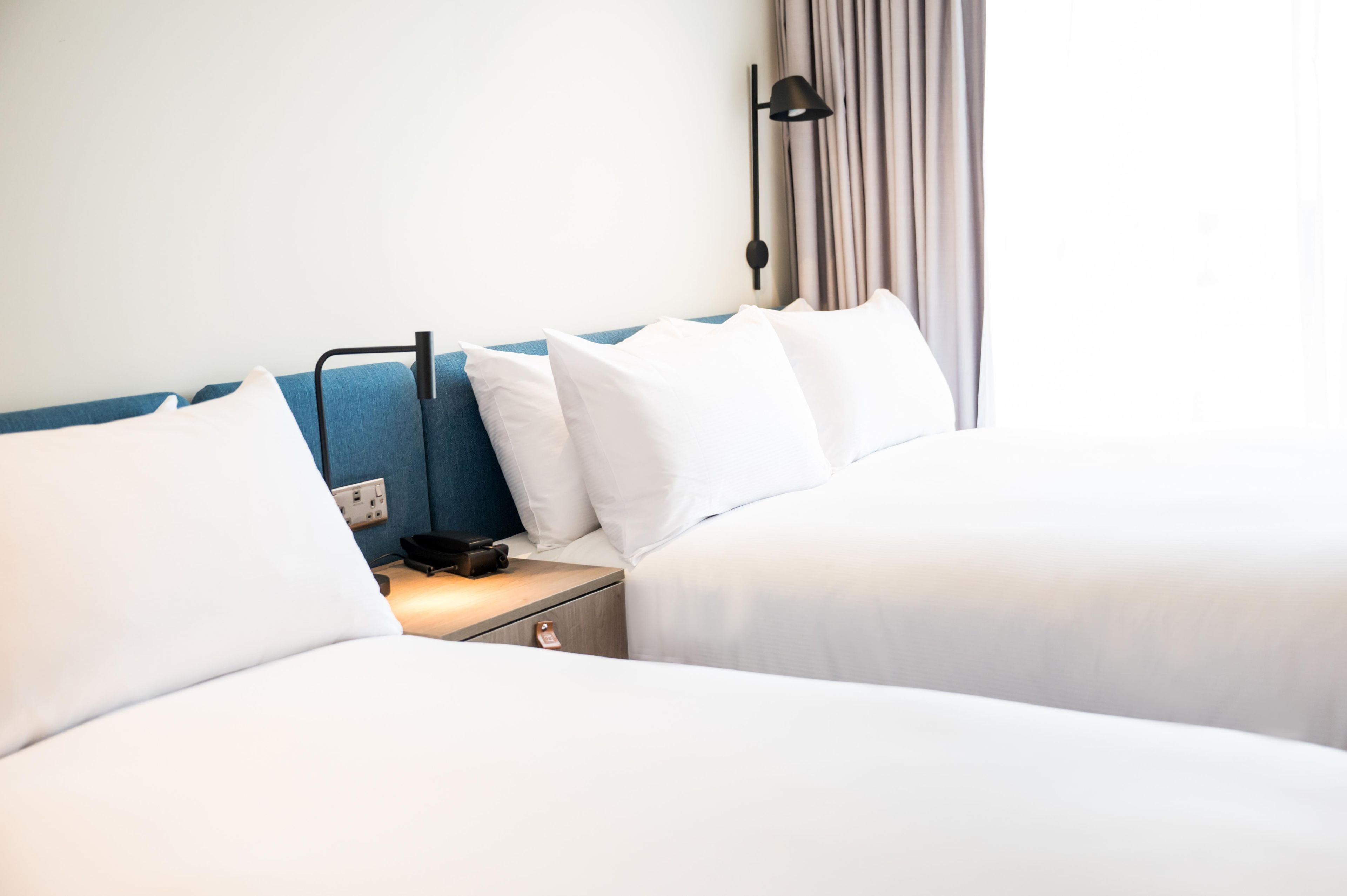 A hotel room with two beds, a bedside table with a lamp, and a power outlet.