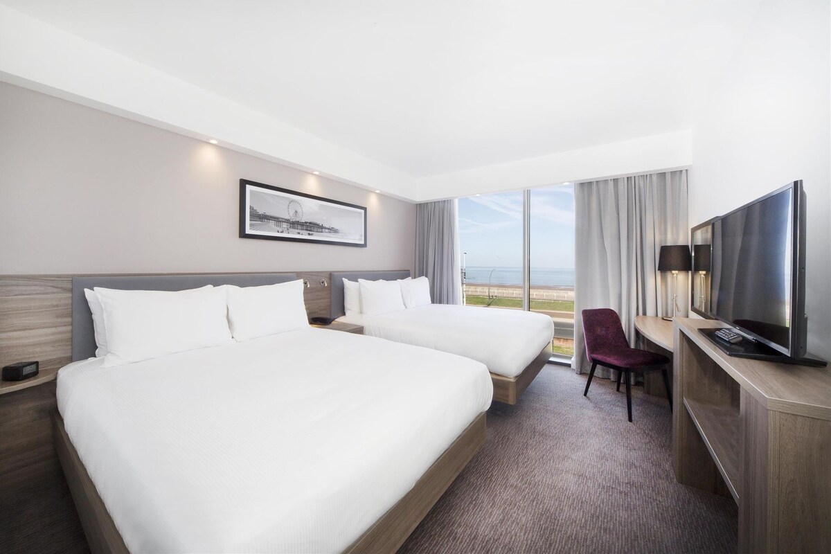 Habitación de hotel con dos camas, un escritorio con televisor y vista a la playa.