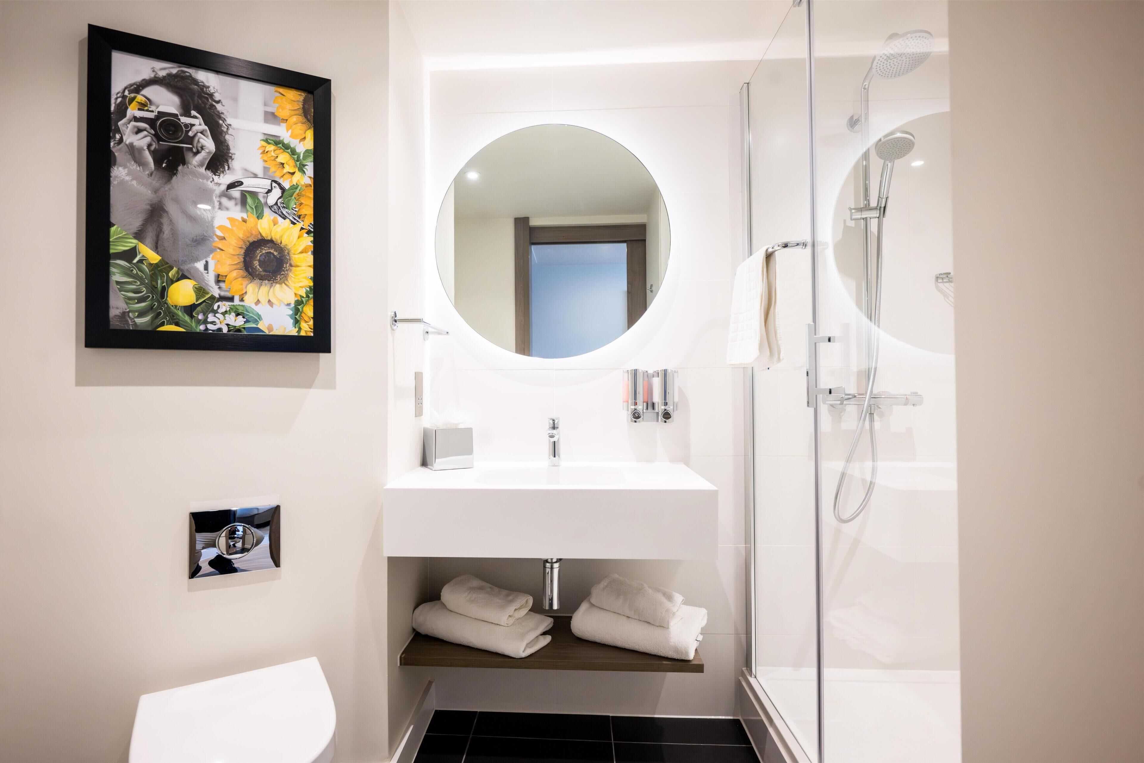 Un baño moderno con un espejo redondo, un lavabo blanco, cabina de ducha con mampara de vidrio y una obra de arte enmarcada en la pared.