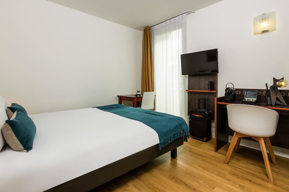A hotel room with a bed, a desk with a computer, a chair, a TV, and a small plant.