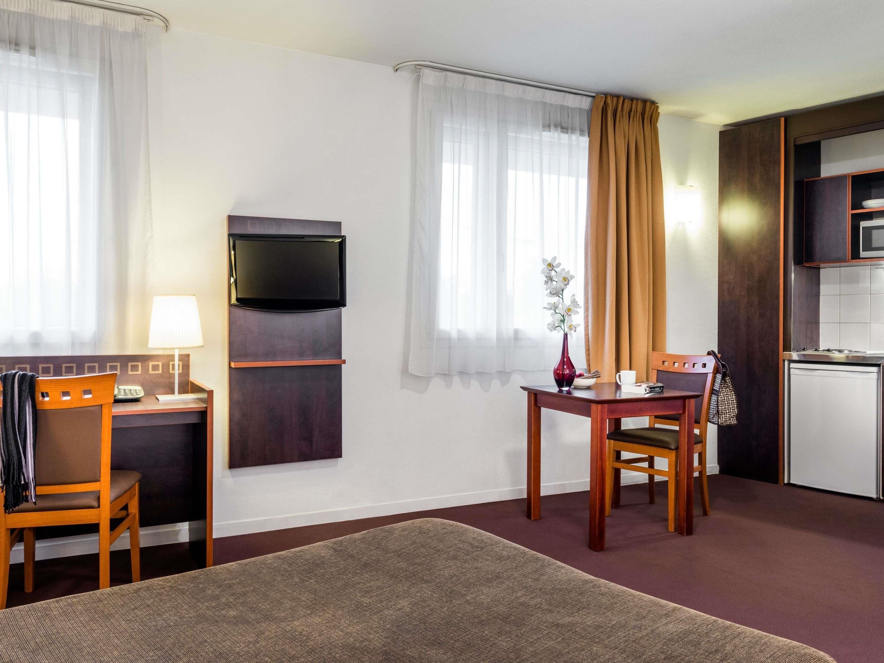 Hotellihuone, jossa on sänky, työpöytä tuolilla, televisio, pieni pöytä maljakolla ja pieni keittiö.