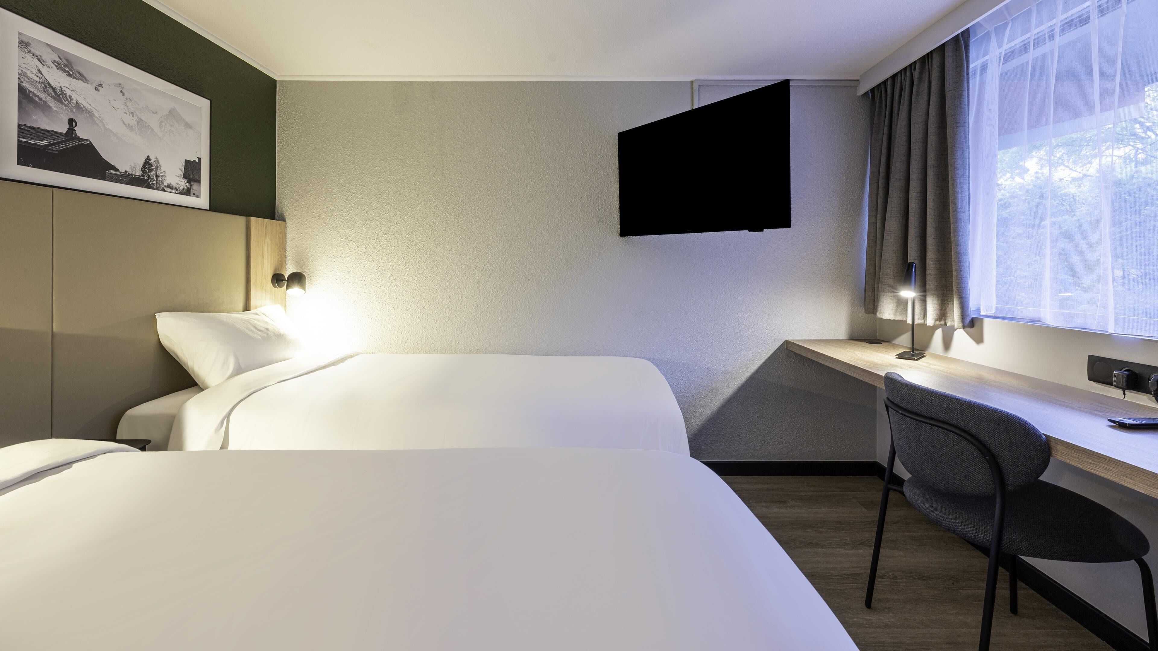 Quarto de hotel com duas camas, uma escrivaninha, uma cadeira, televisão presa à parede e janela com cortinas.