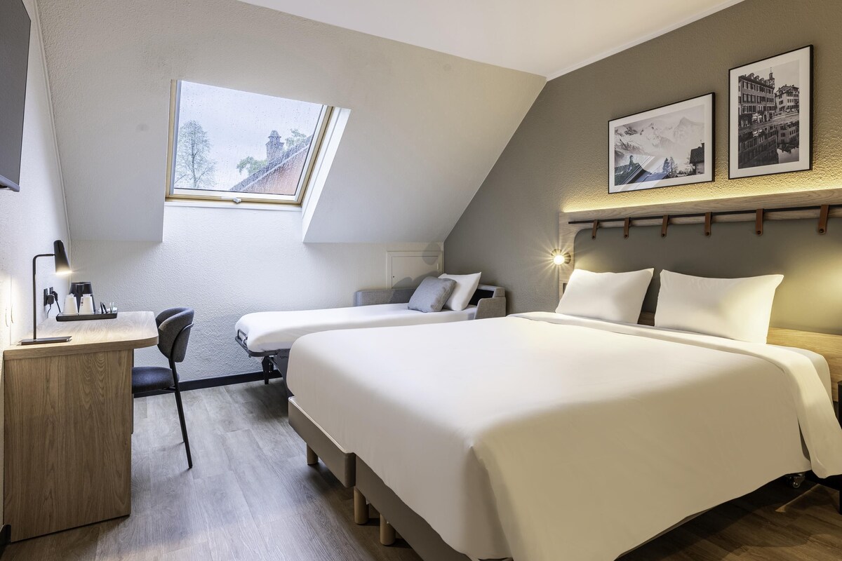 Una habitación de hotel moderna con dos camas, un escritorio con lámpara, una silla y una ventana con vistas a árboles.