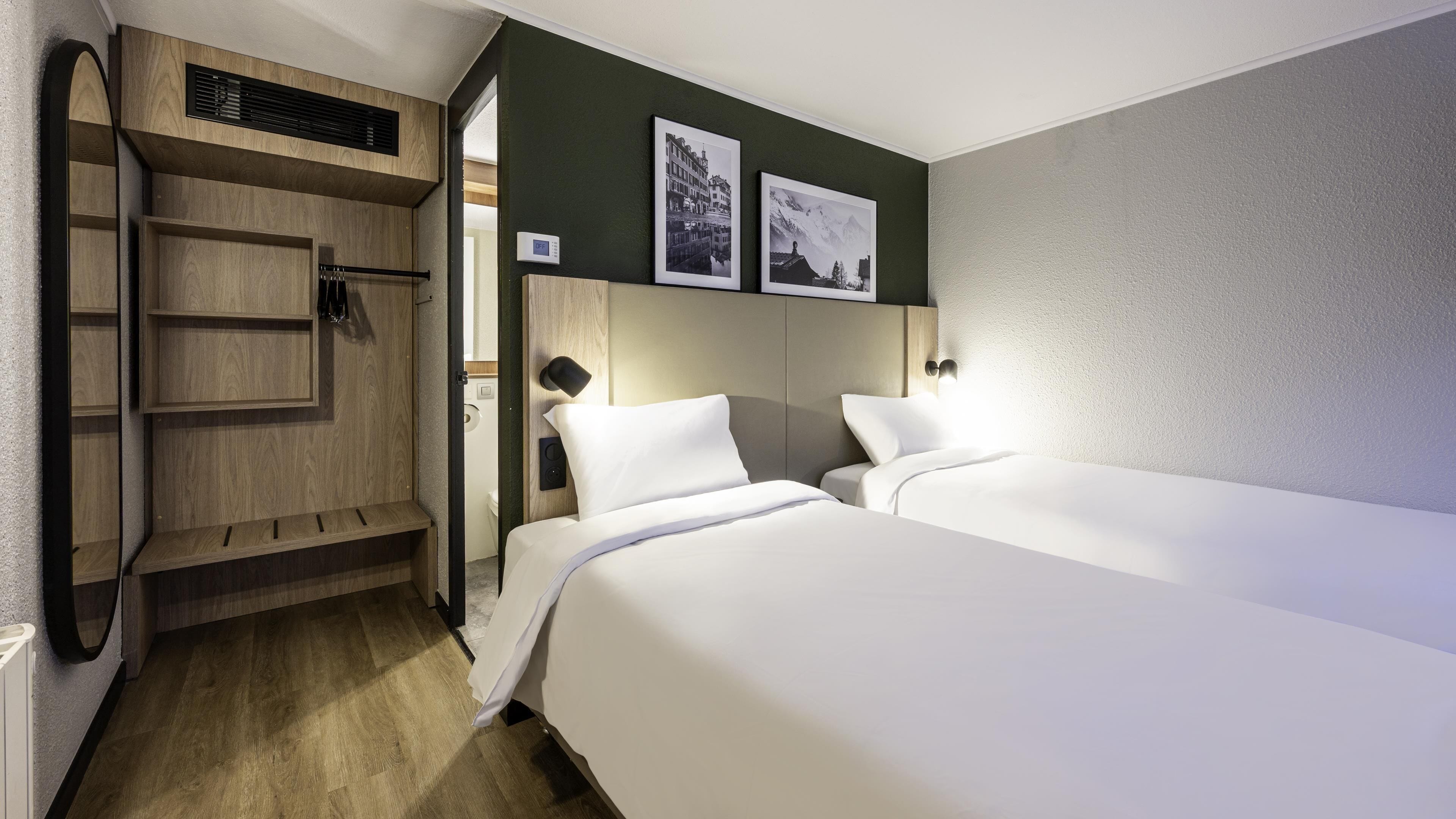 A hotel room with two beds, a wooden wardrobe, a built-in heater, and two framed pictures on the wall.