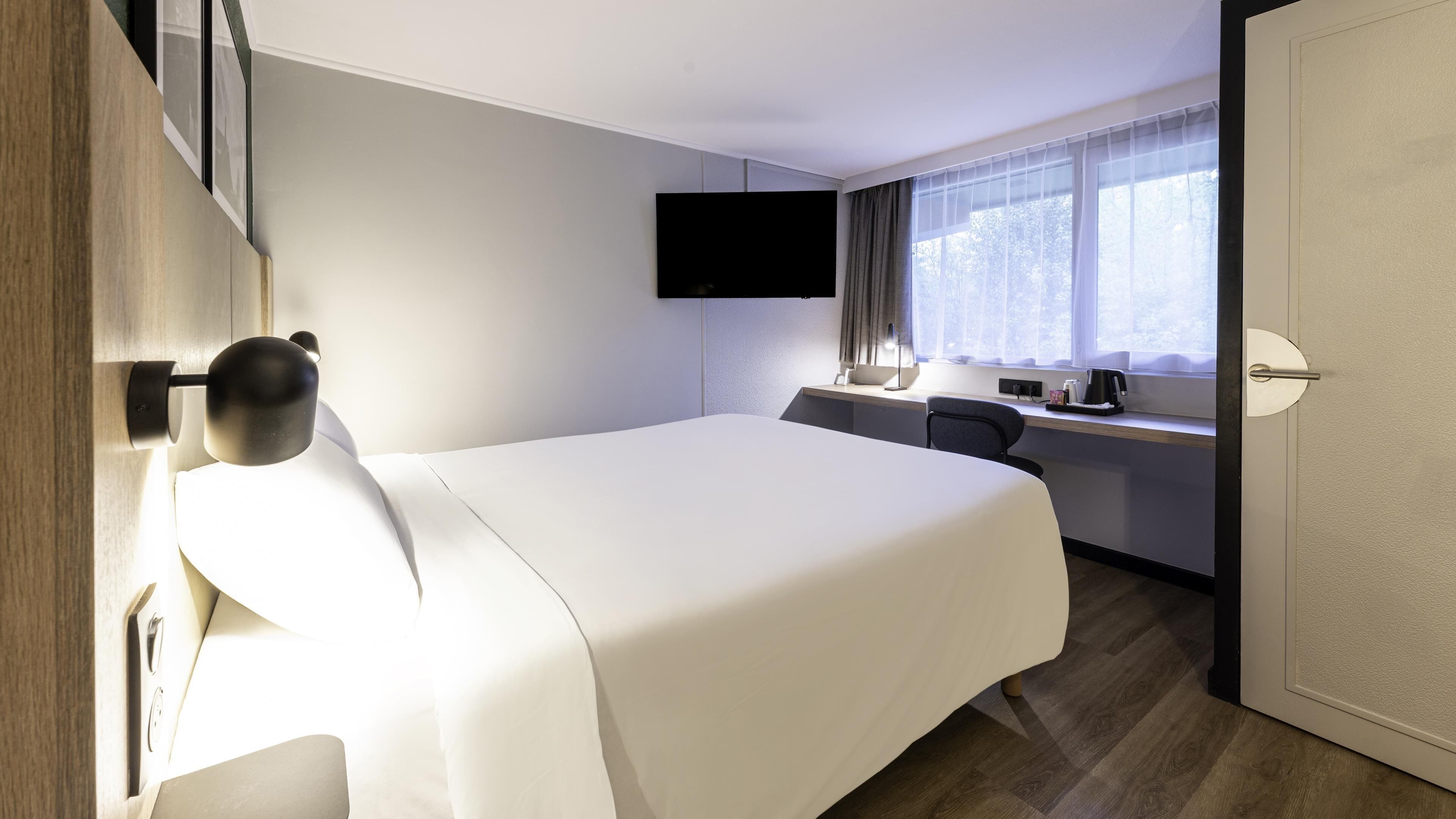 Quarto de hotel com cama, escrivaninha, cadeira, televisão fixada na parede e janela com cortinas.