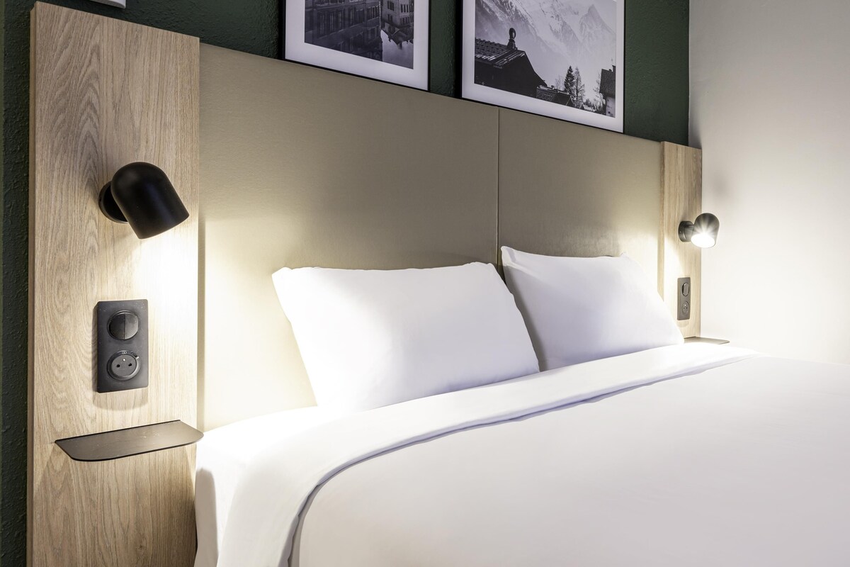 Una cama bien tendida con sábanas blancas, un cabecero de madera y dos lámparas de lectura fijadas a la pared.