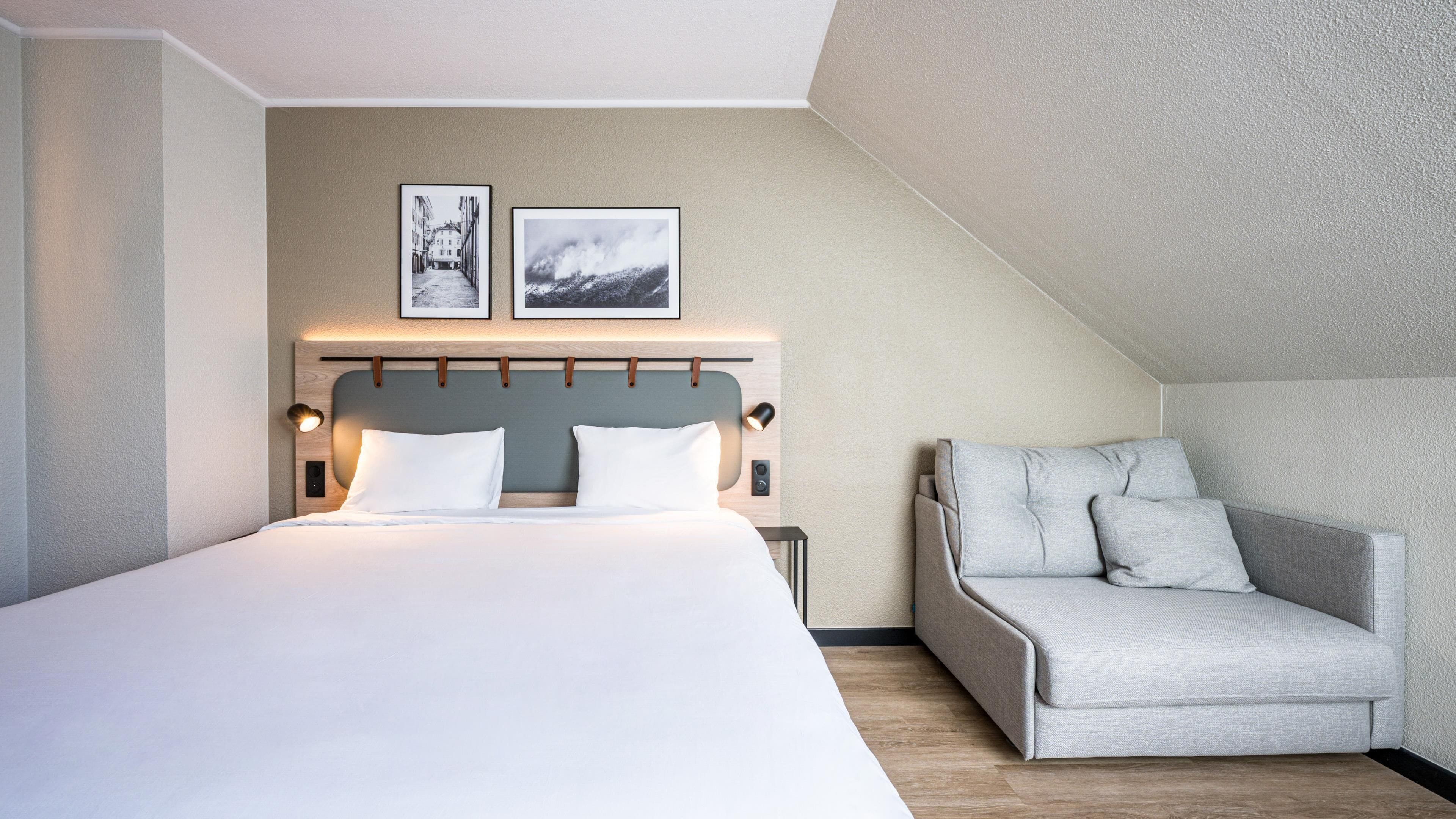 A modern hotel room with a large bed, a sofa, two framed pictures on the wall, and a sloped ceiling.