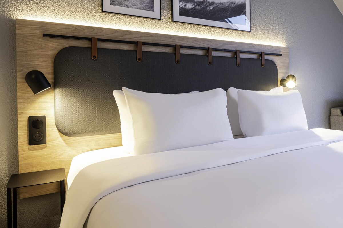 Una habitación de hotel moderna con una cama bien hecha, cabecera de madera y una lámpara fijada a la pared.