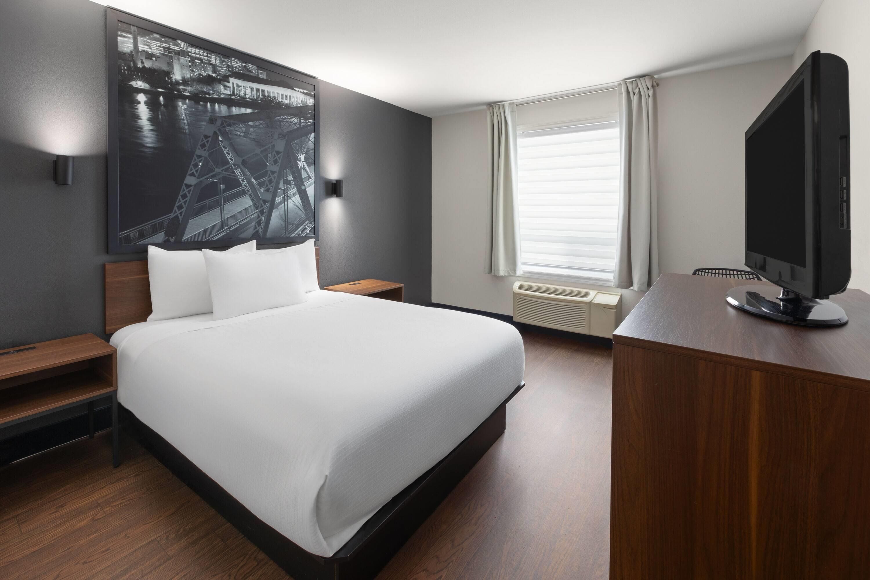 A modern hotel room with a large bed, a desk with a flat-screen TV, a wall-mounted TV, a window with curtains, and a cityscape painting on the wall.