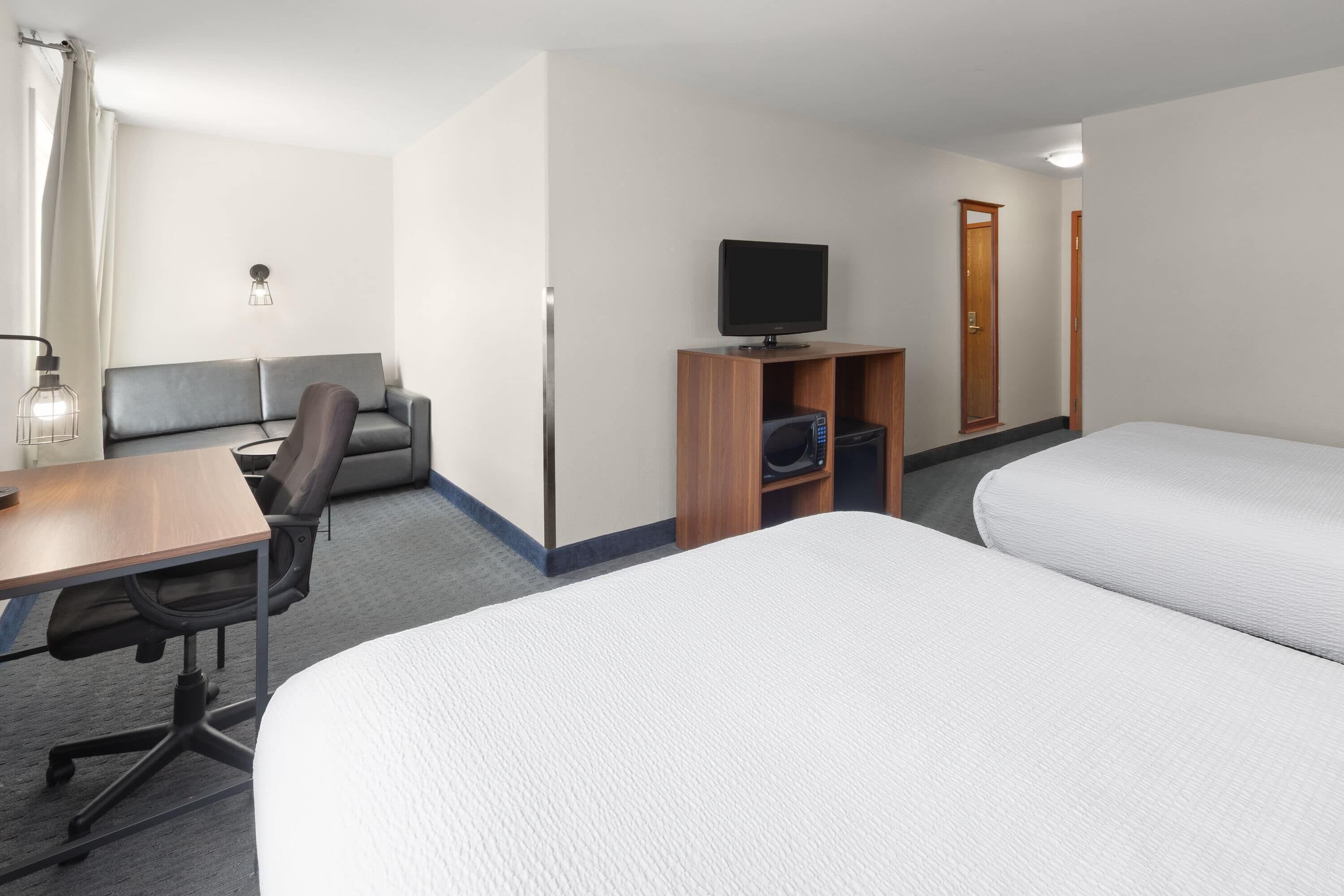 Habitación de hotel con dos camas, un escritorio con una silla, un televisor sobre un soporte de madera y una pequeña zona de estar con un sofá y una silla.