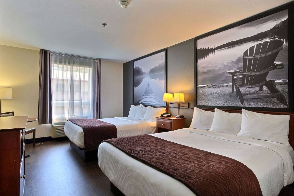 A hotel room with two beds, a desk, a chair, a nightstand with a lamp, and a large framed picture on the wall.