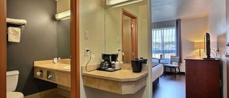Een hotelbadkamer met toilet, wastafel en spiegel. Op het aanrecht staat een koffiezetapparaat en een prullenbak. De kamer heeft een bed en een stoel.