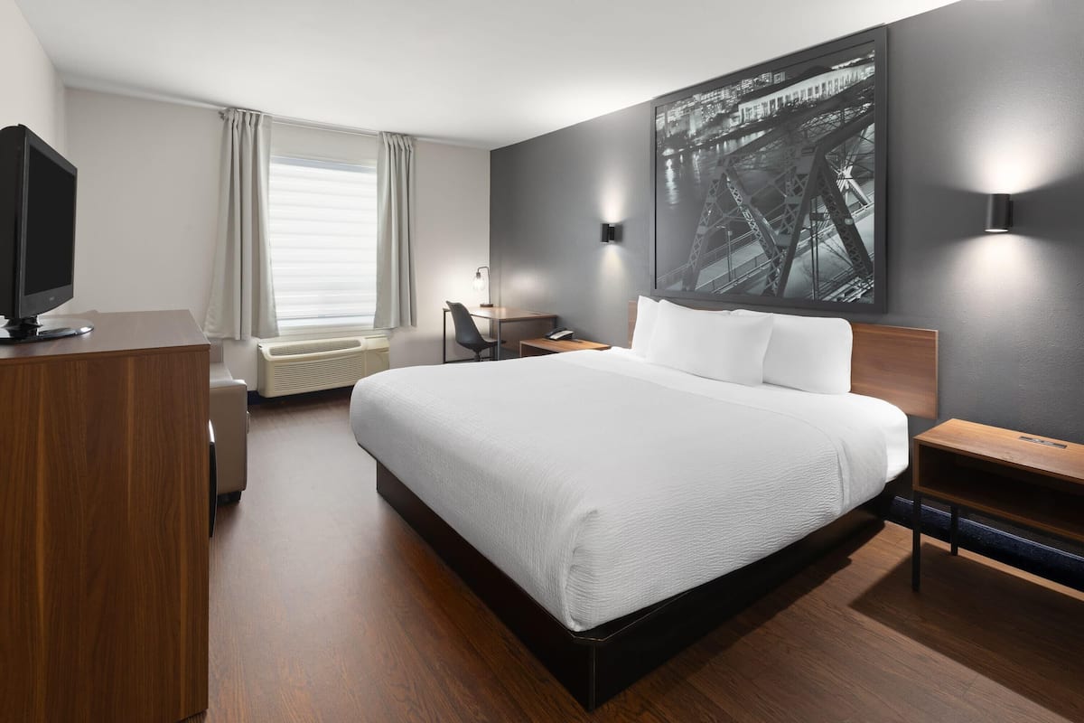 A modern hotel room with a large bed, a desk, a chair, a television, and a large window with curtains.