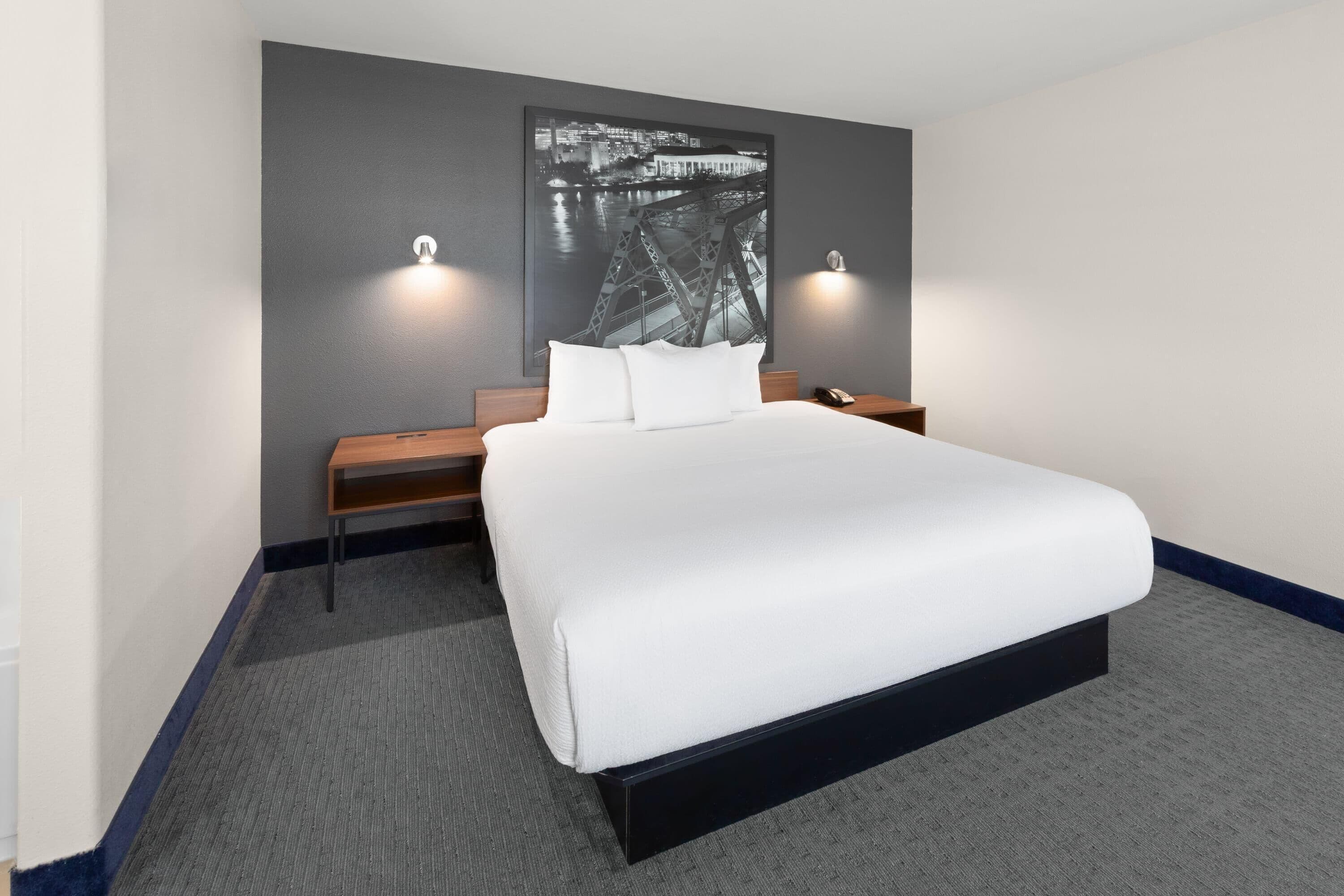 A hotel room with a large bed, a nightstand, a desk, and a wall-mounted picture of a bridge.