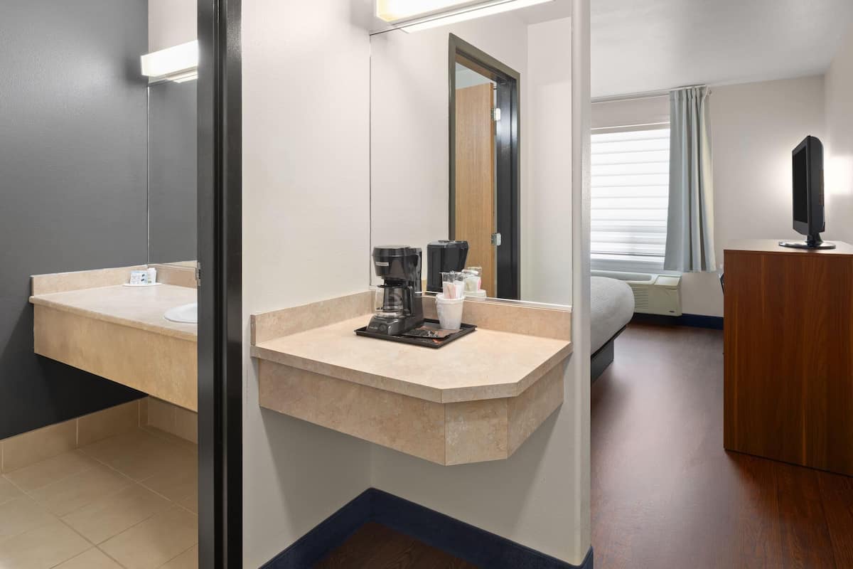 A hotel room with a curved sink, a coffee maker, a mirror, a bed, a TV, and a window with curtains.