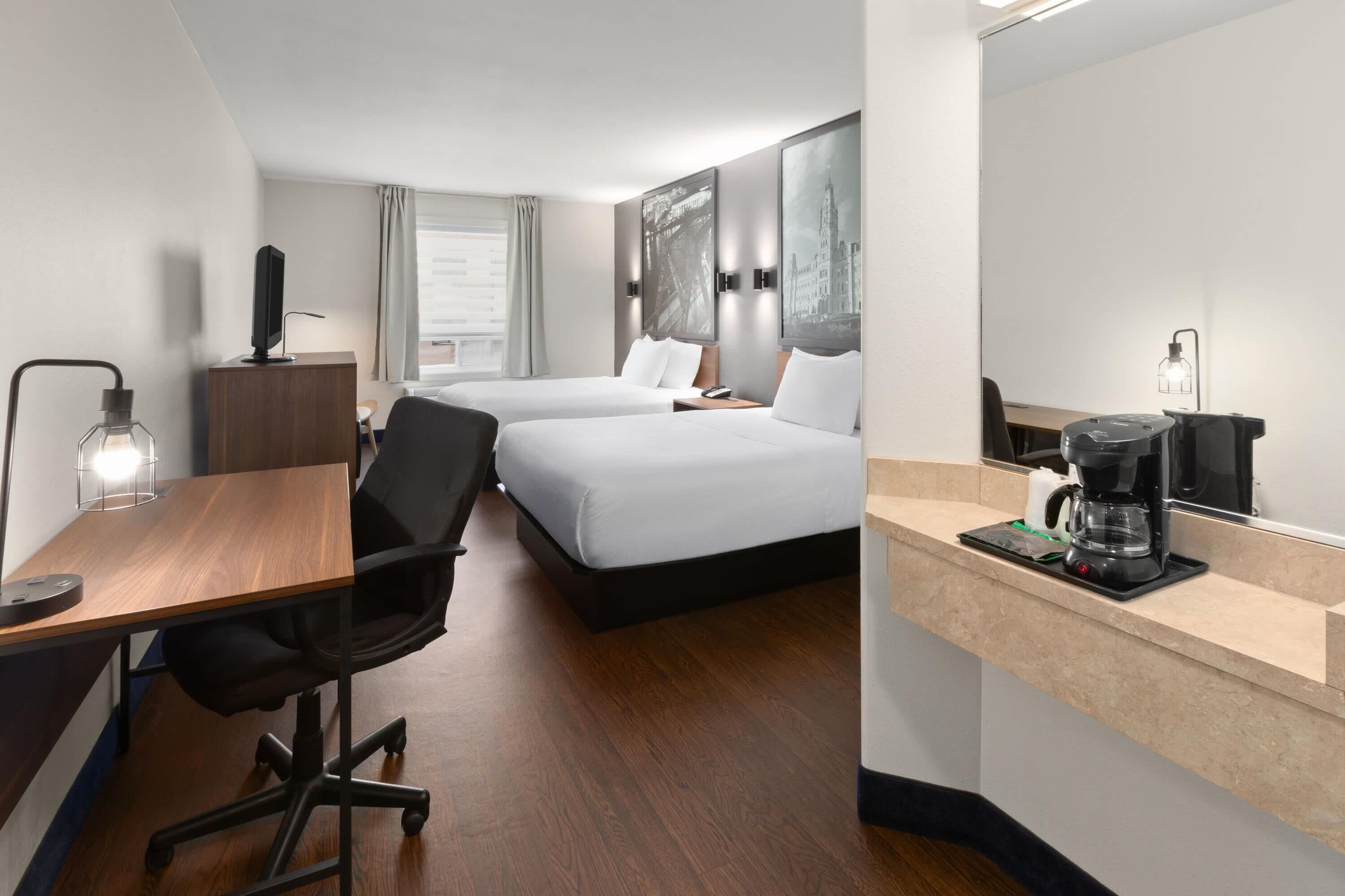 A hotel room with a wooden desk, two beds, a coffee machine, and a large mirror.