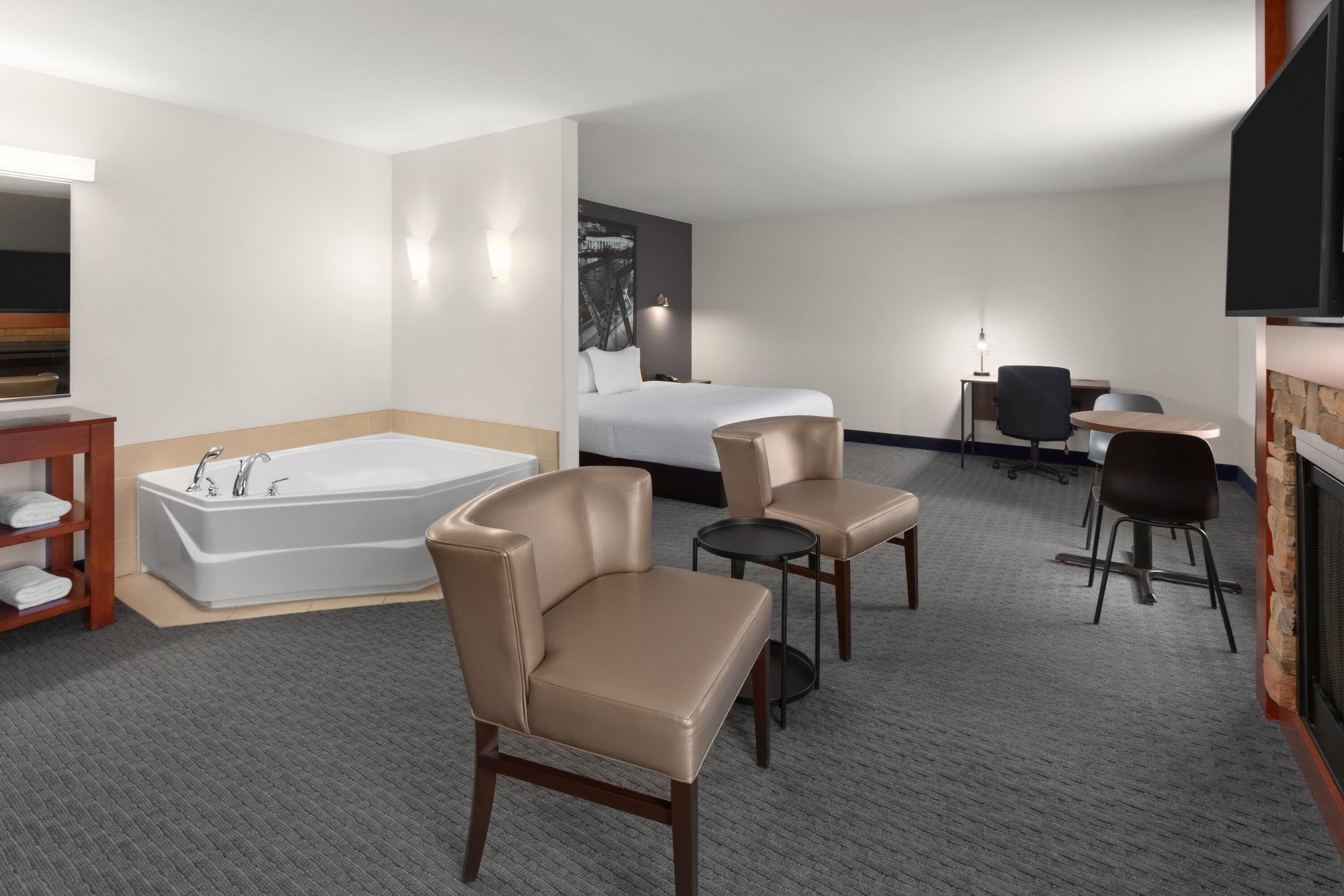 Une chambre d’hôtel avec une grande baignoire, deux fauteuils beiges, un lit, un bureau avec une chaise et un téléviseur fixé au mur.