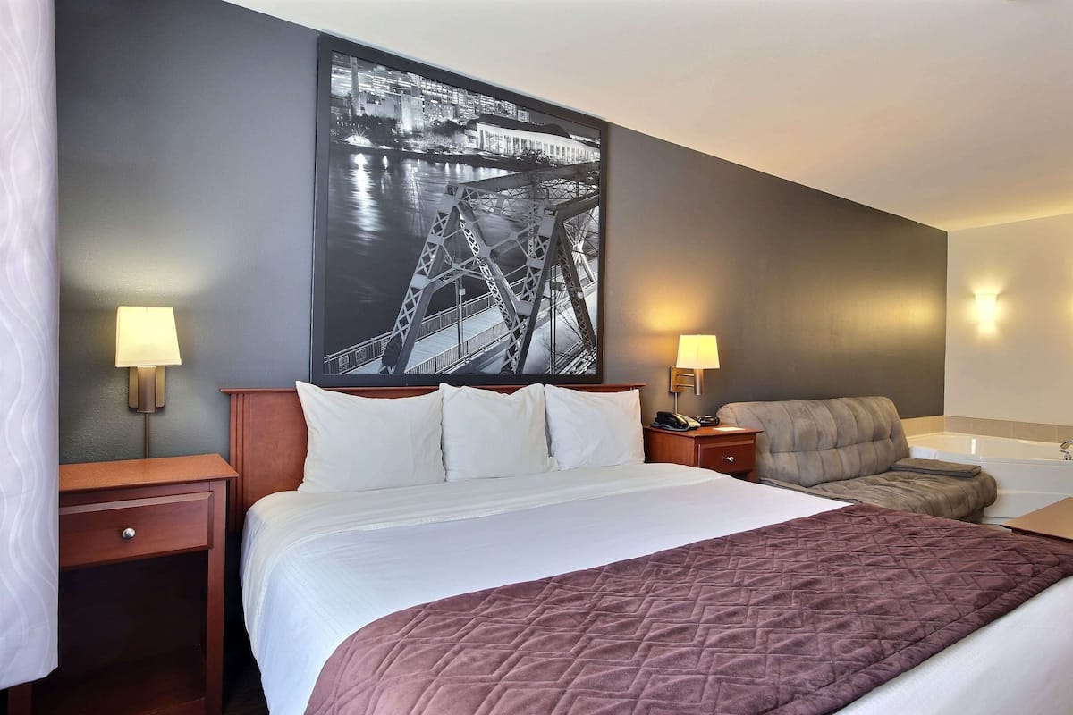A hotel room with a large bed, a nightstand, a lamp, a sofa, and a wall-mounted picture of a bridge.
