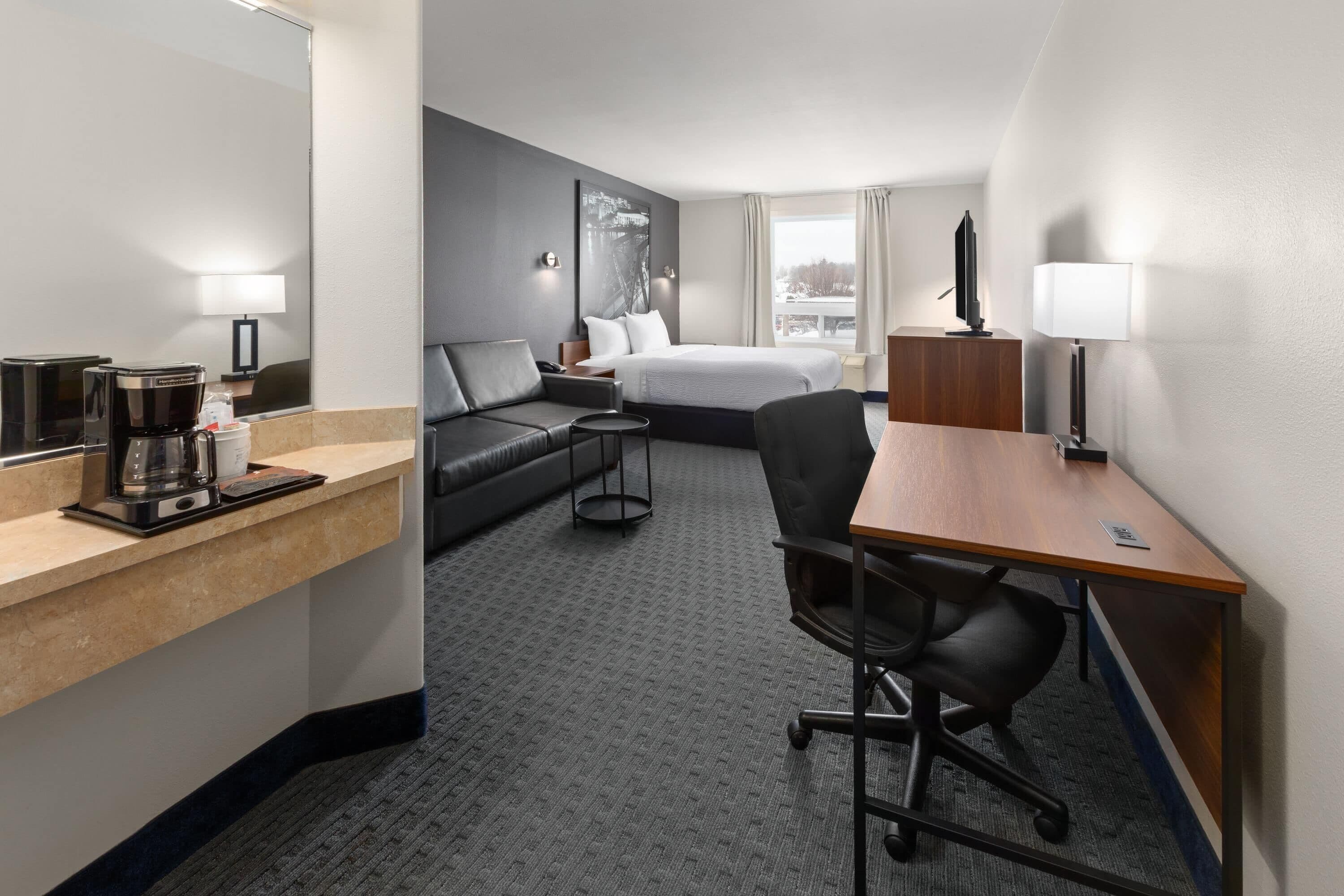 A hotel room with a bed, a desk, a chair, a sofa, a coffee maker, and a TV.