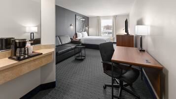 A hotel room with a bed, a desk, a chair, a sofa, a coffee maker, and a TV.