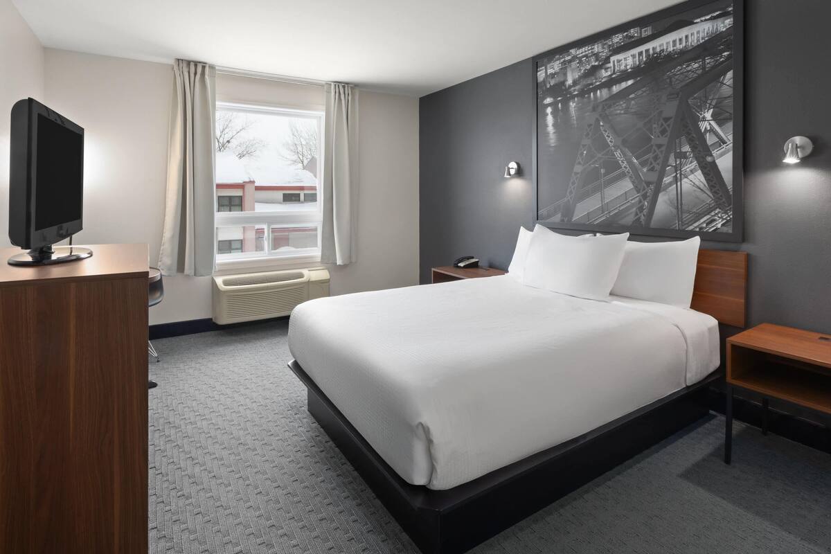 A hotel room with a large bed, a flat-screen TV, a window with curtains, a wall-mounted light, and a framed picture of a bridge.