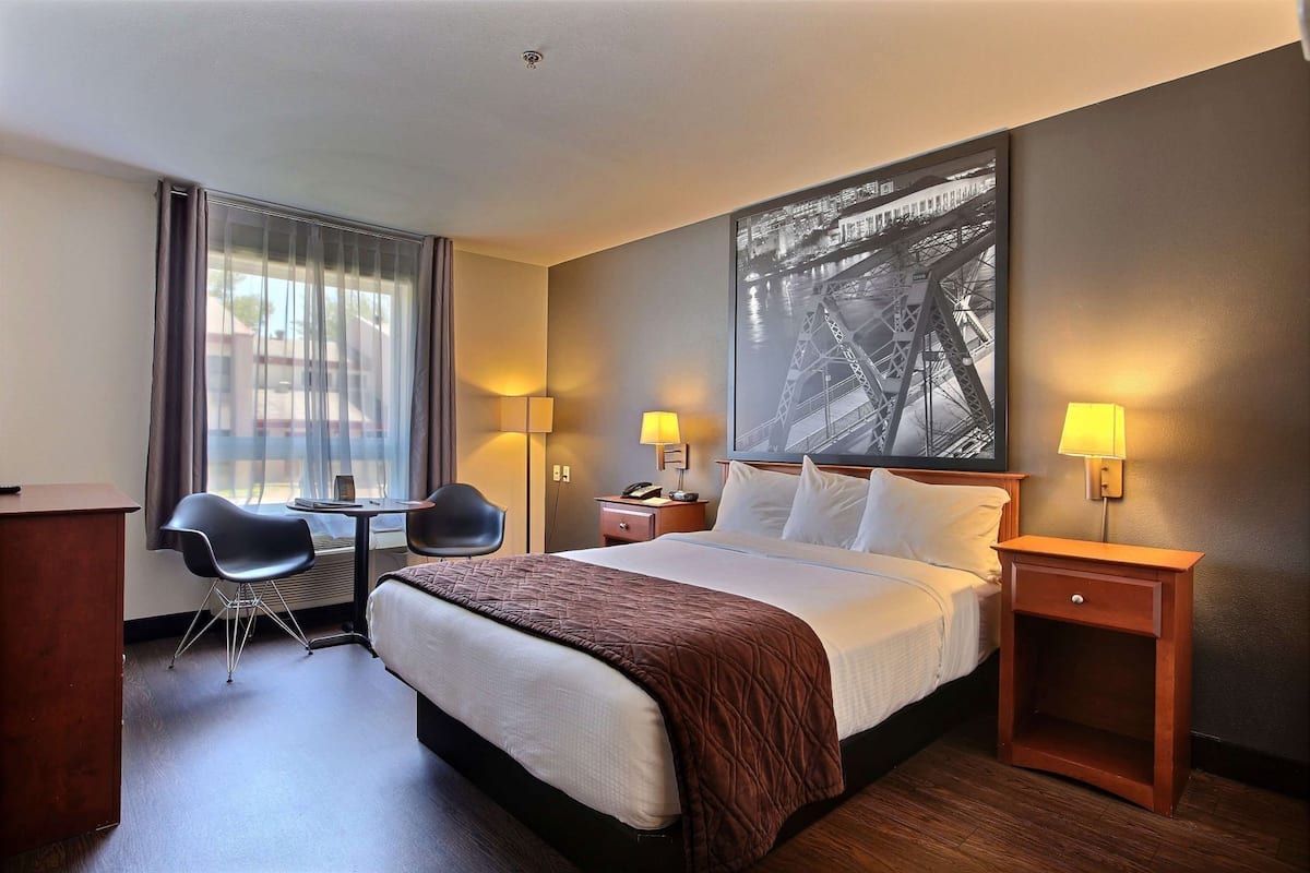 A hotel room with a bed, two bedside tables, a desk with a chair, a large window with curtains, and a wall-mounted lamp.