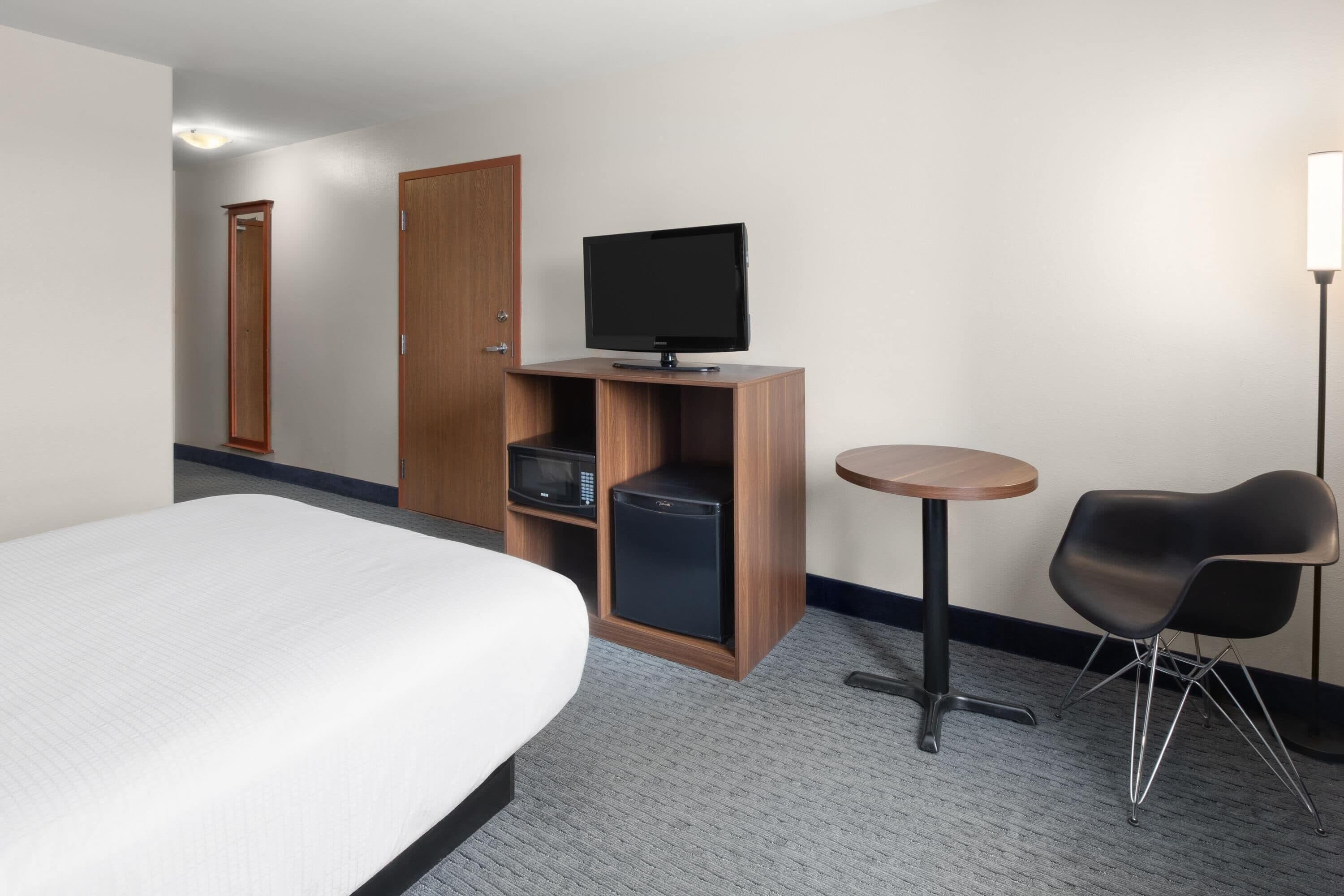 A hotel room with a bed, a television on a wooden stand, a small refrigerator, a microwave, a round table with a chair, and a door leading to another room.