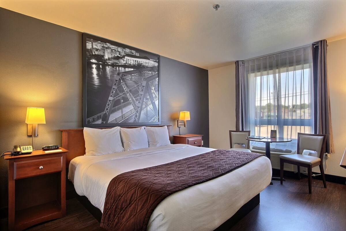 A hotel room with a large bed, two bedside tables with lamps, a desk with a chair, and a large window with curtains.