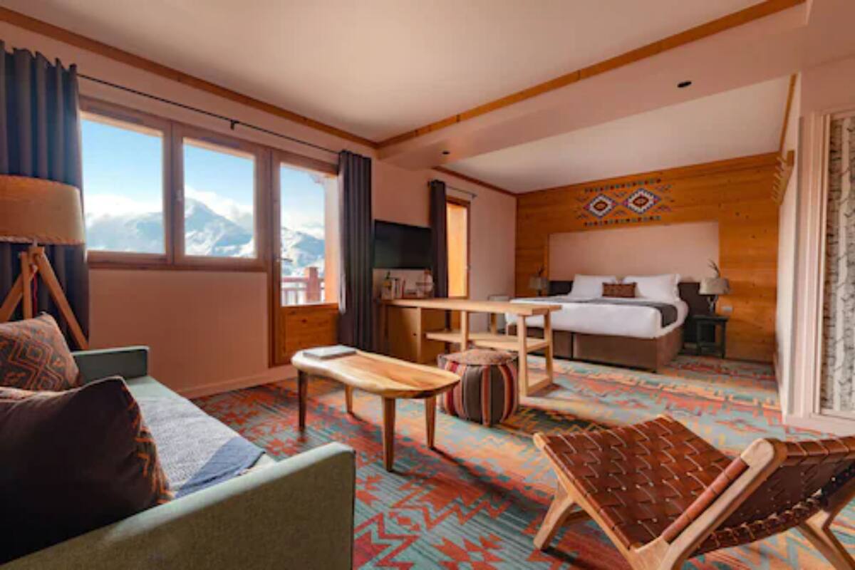 A hotel room with a bed, a desk, a chair, a sofa, and a view of mountains.