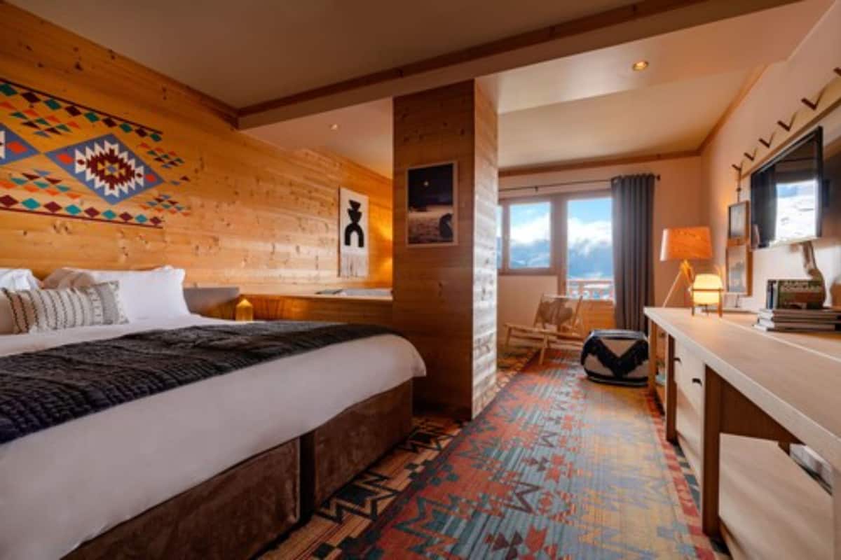 A hotel room with a large bed, a wooden headboard, a colorful rug, a desk with a lamp, and a view of the mountains through the window.