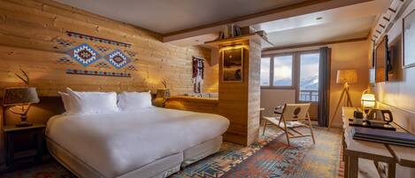 Une chambre d’hôtel avec un grand lit, une tête de lit en bois, un tapis à motifs, un bureau avec une lampe et une fenêtre donnant sur la montagne.